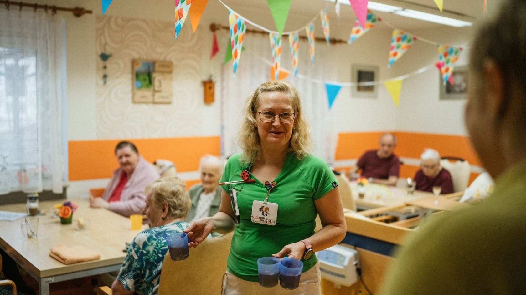 Carer offering drinks to residents in a care home lounge decorated with bunting