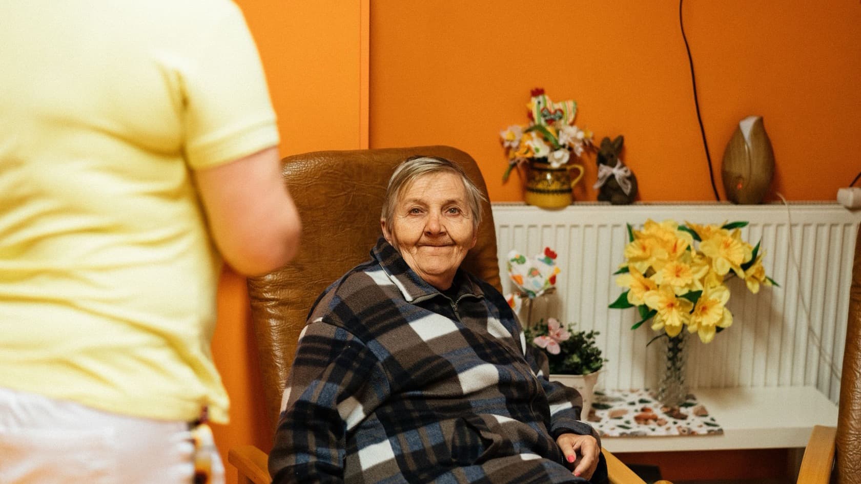 Resident sitting alone in an armchair in her room while a carer stands nearby