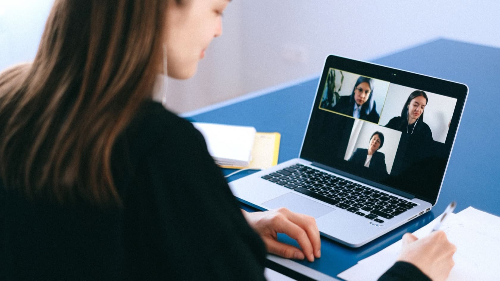 Woman taking notes by hand during a video call with three colleagues on her laptop