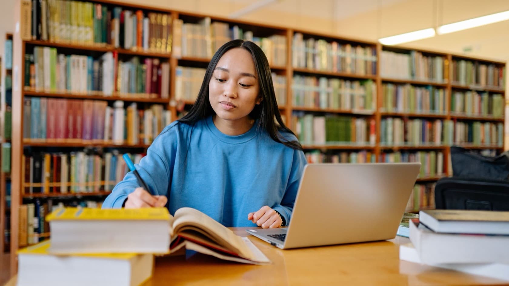 Student taking notes from a textbook beside her laptop in a library