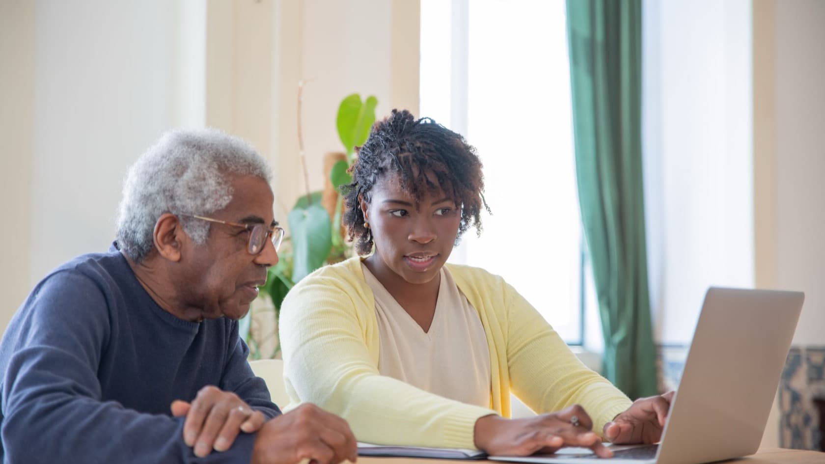 Care worker typing up a form on a laptop while an older resident sits beside her at a table