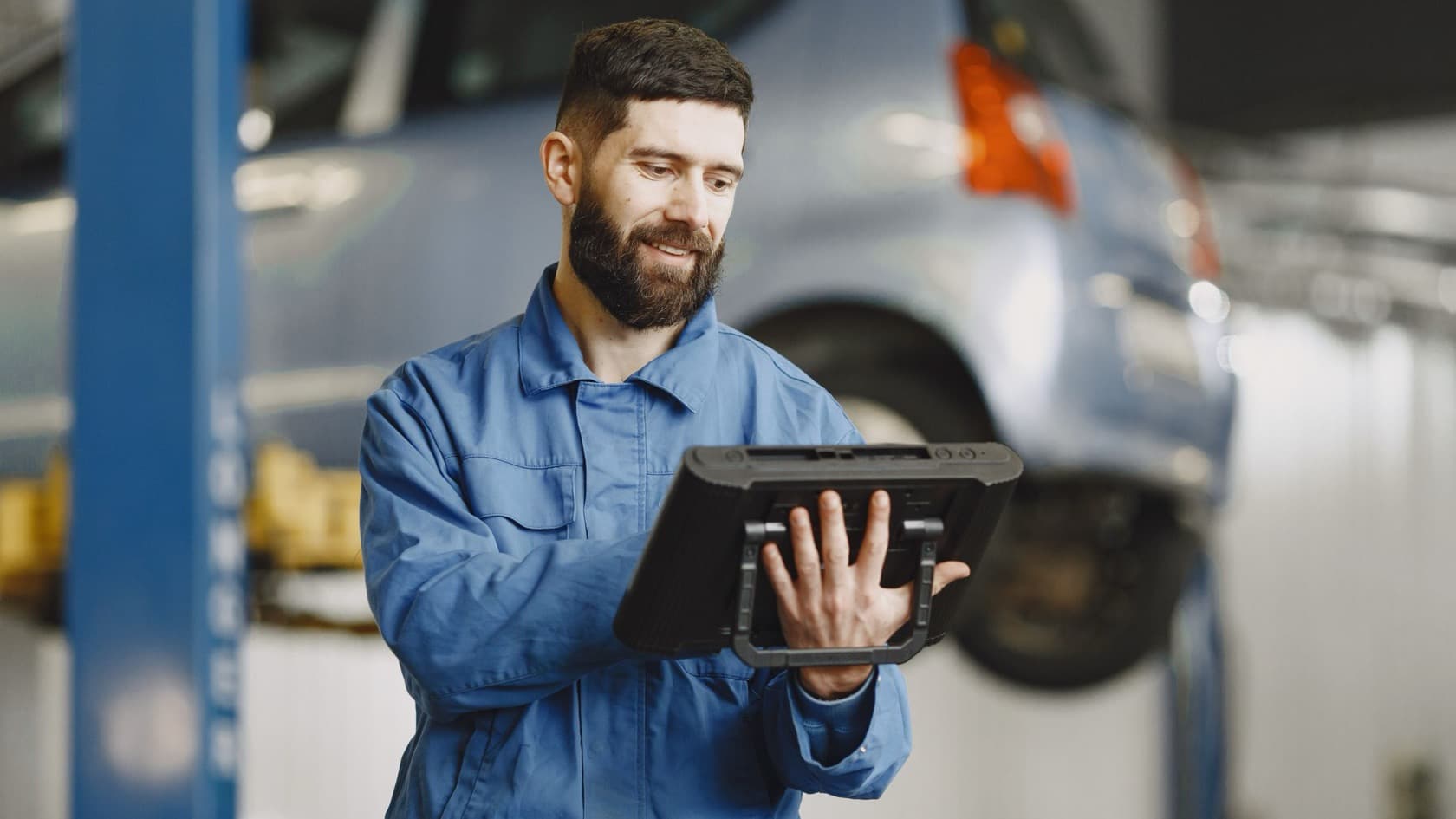 Mechanic in overalls looking something up on a rugged tablet in a garage with a car on a lift