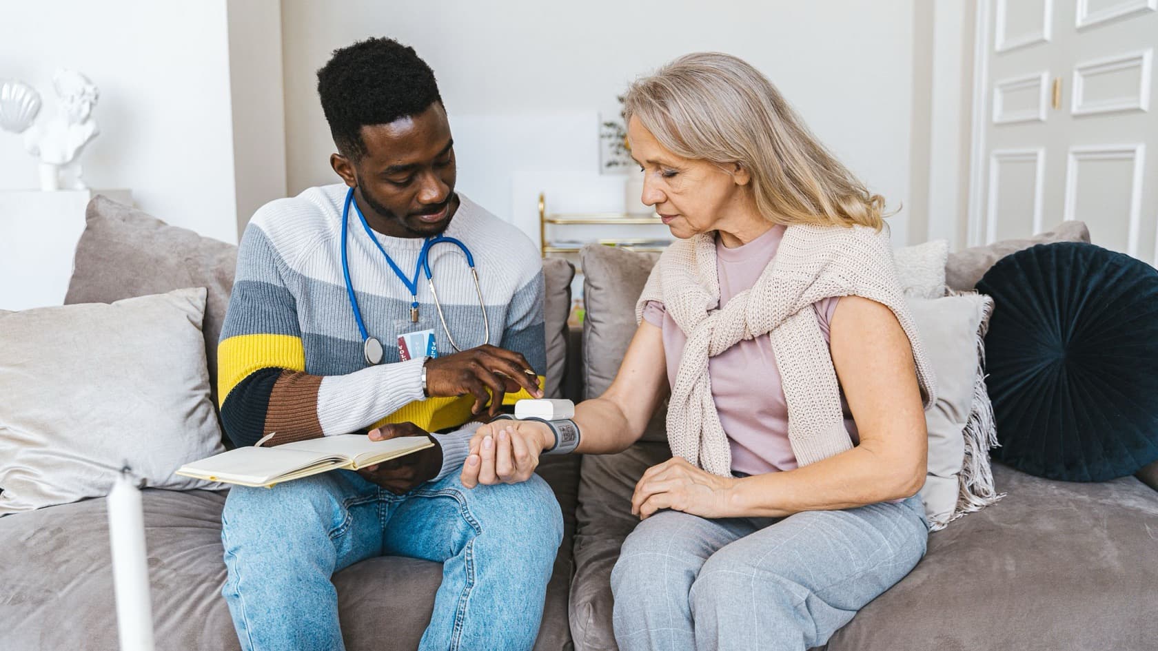 Carer taking an older woman's blood pressure on the sofa with a notebook open on his knee