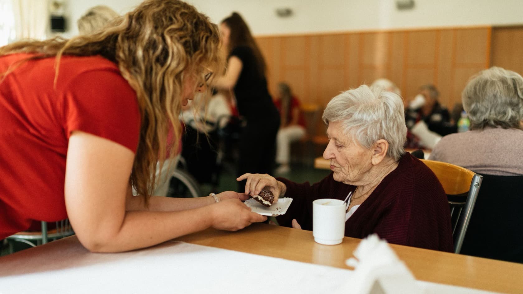Carer serving an older resident a snack at a dining table in a care home lounge