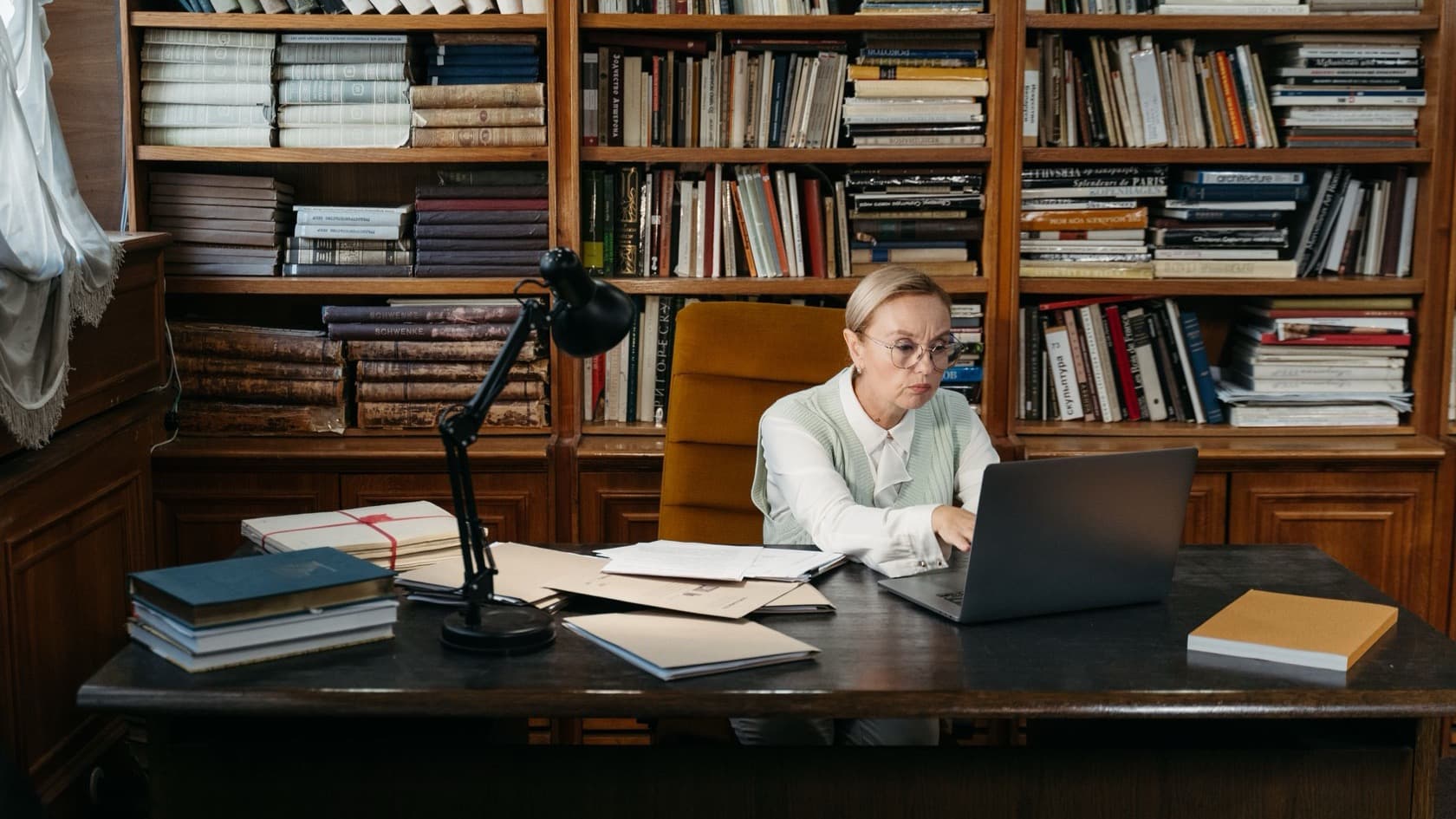 Publisher working on a laptop at a desk of manuscripts and books in front of full bookshelves