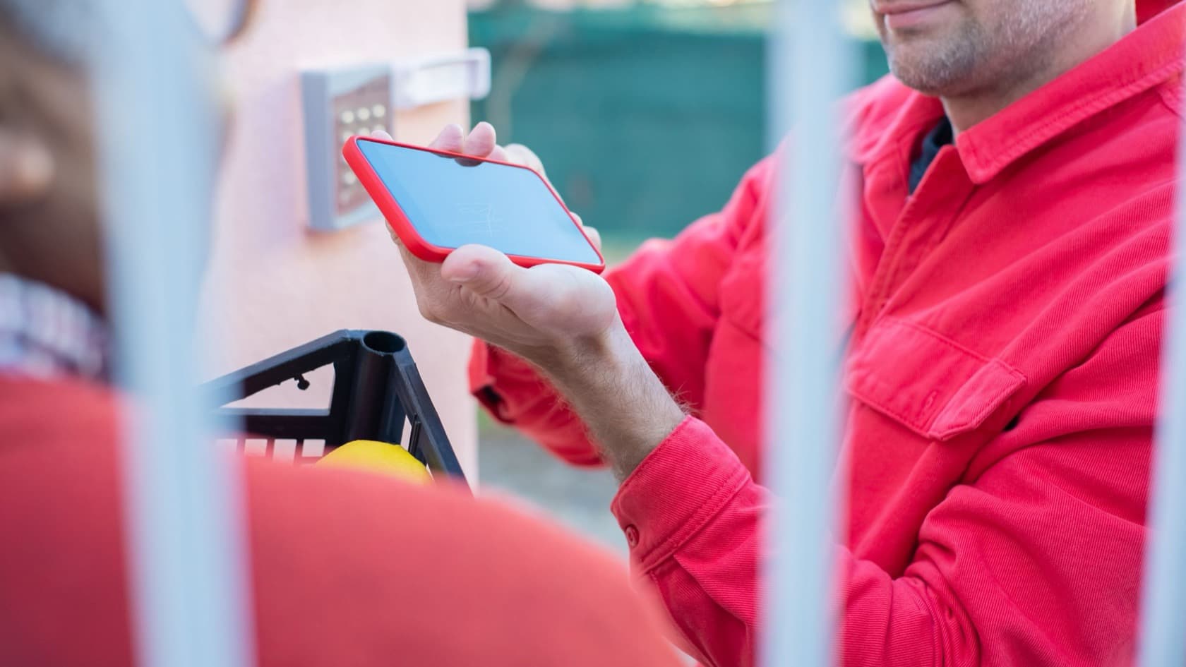 Visitor holding up a phone pass at a gated entrance with a keypad, seen through the gate bars