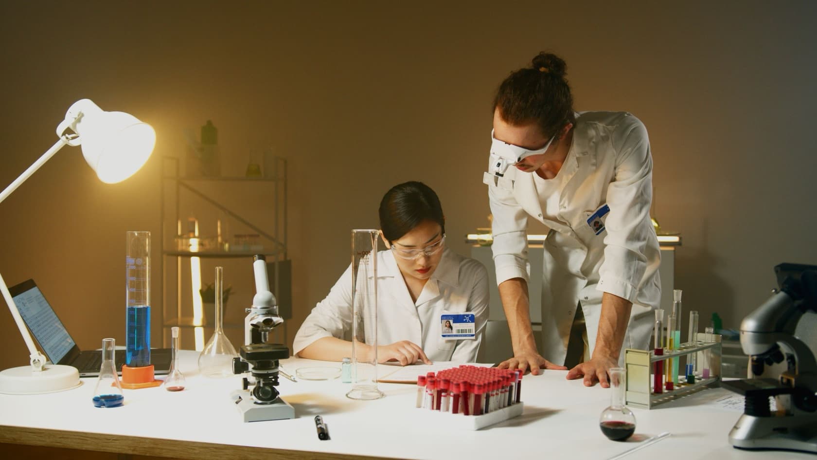 Two lab scientists reviewing study notes at a bench with sample tubes and microscopes