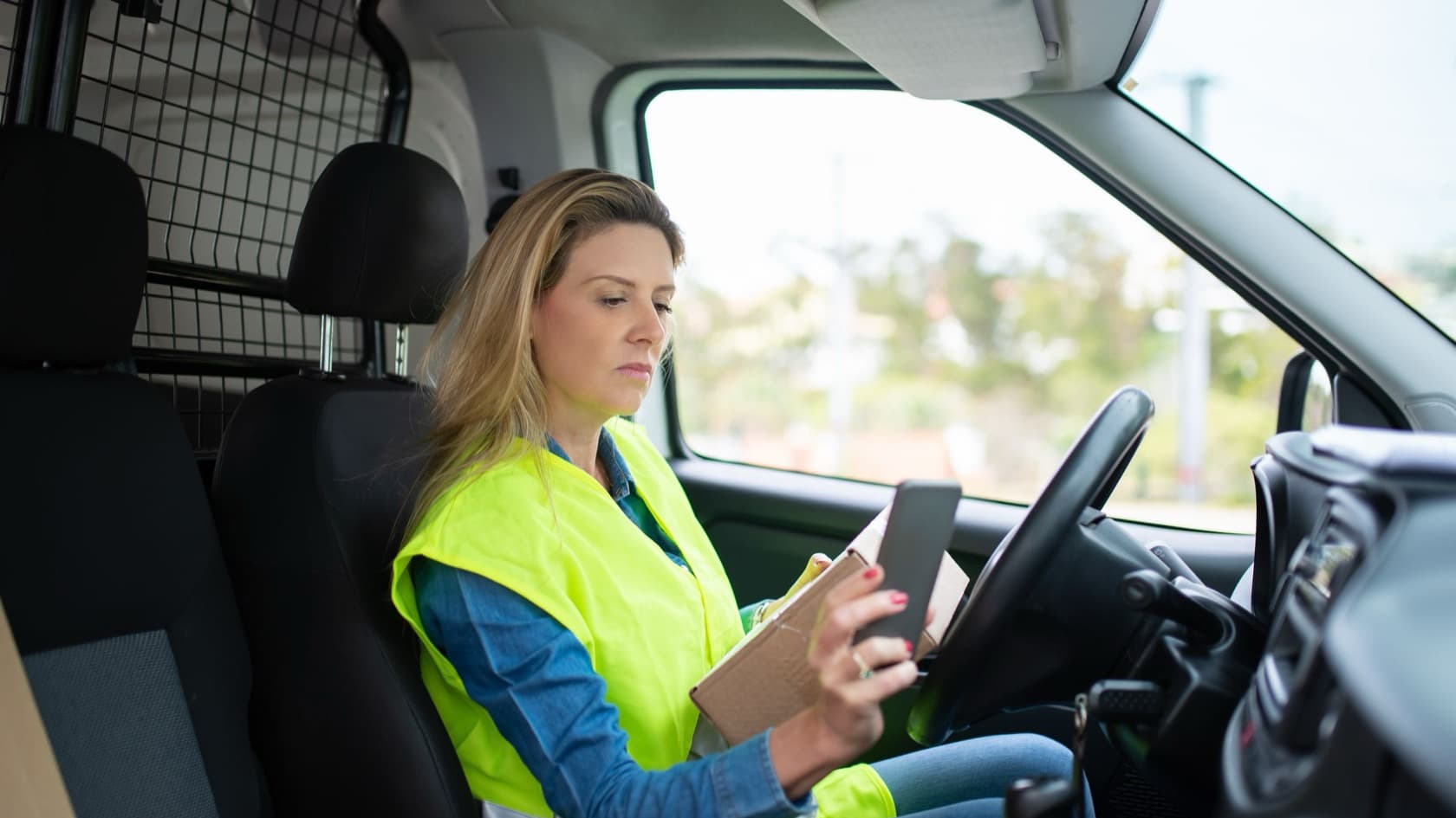 Field worker in a high-visibility vest checking her phone and a parcel in the driver's seat of a van