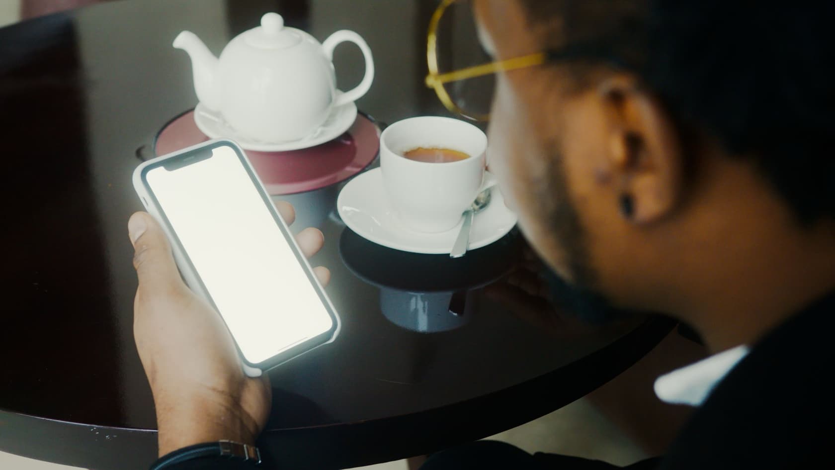 Reader holding a phone over a table with a teapot and a cup of tea, seen over his shoulder