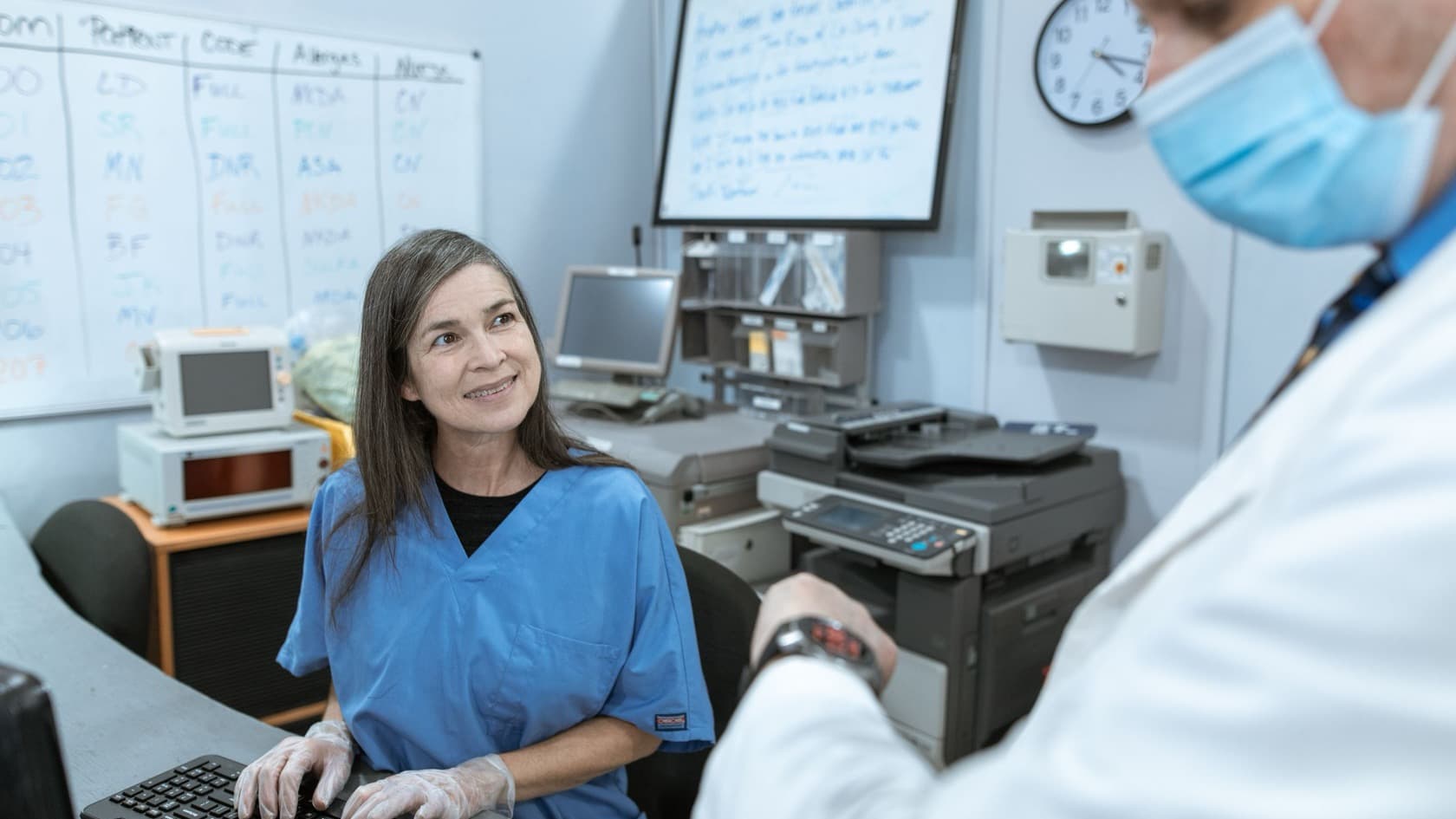 Hospital staff member at an admin desk computer turning to talk with a doctor