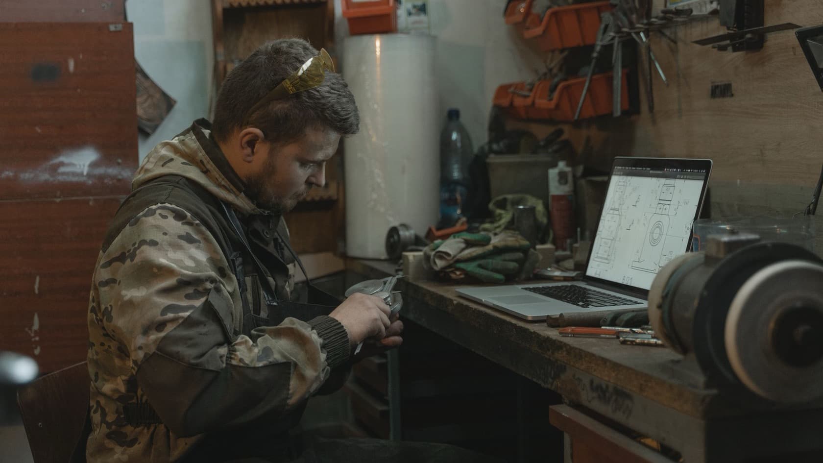 Engineer checking a metal part at a workshop bench beside a laptop showing a technical drawing