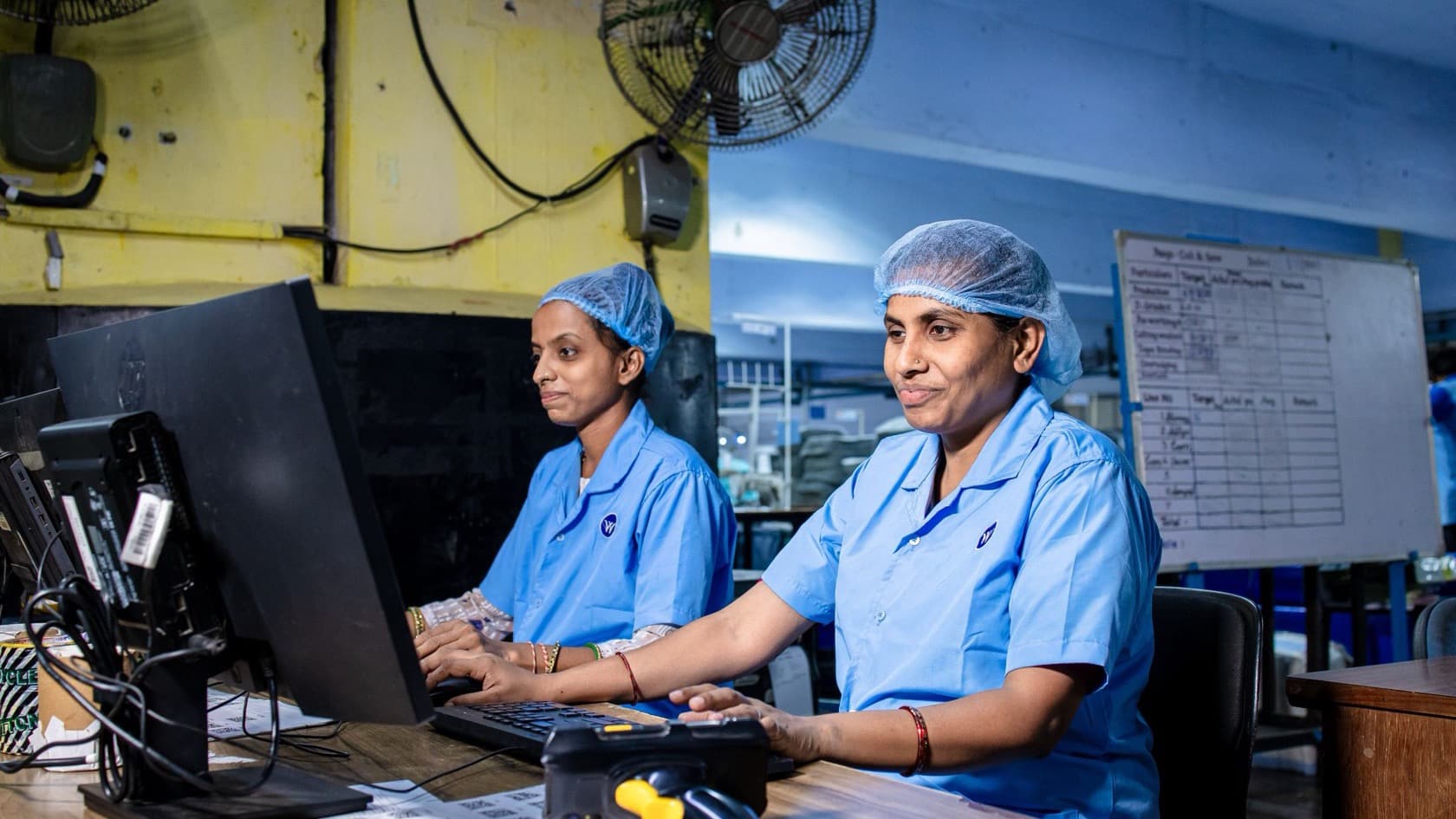 Two factory staff working at computers with a barcode scanner, production whiteboard behind