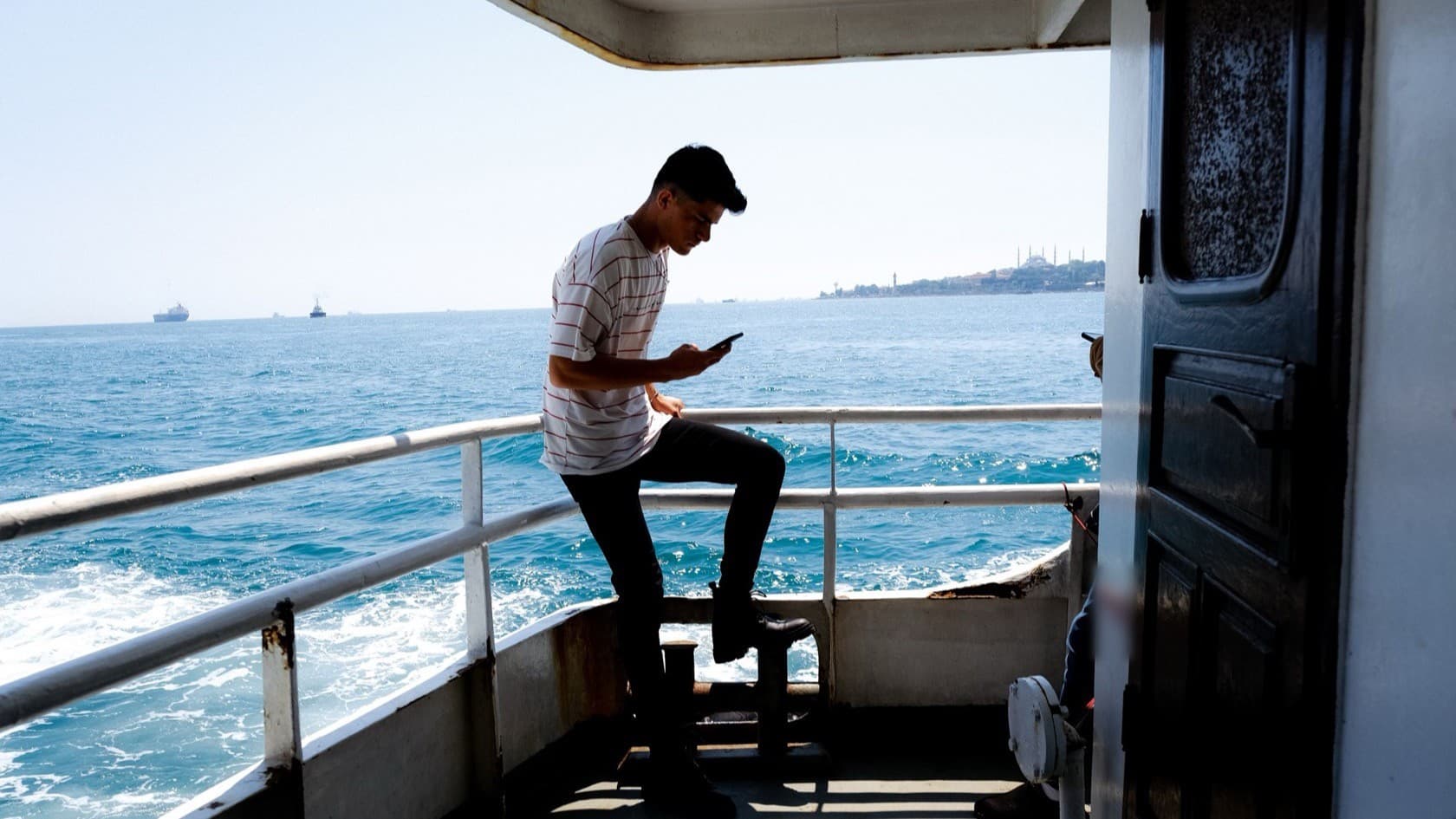 Man reading on his phone at the deck rail of a ship at sea, coastline on the horizon