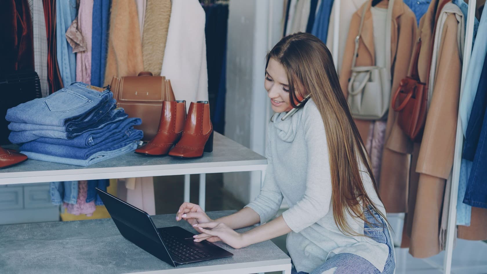 Shop owner updating listings on a laptop among clothing rails and shelved stock in her boutique