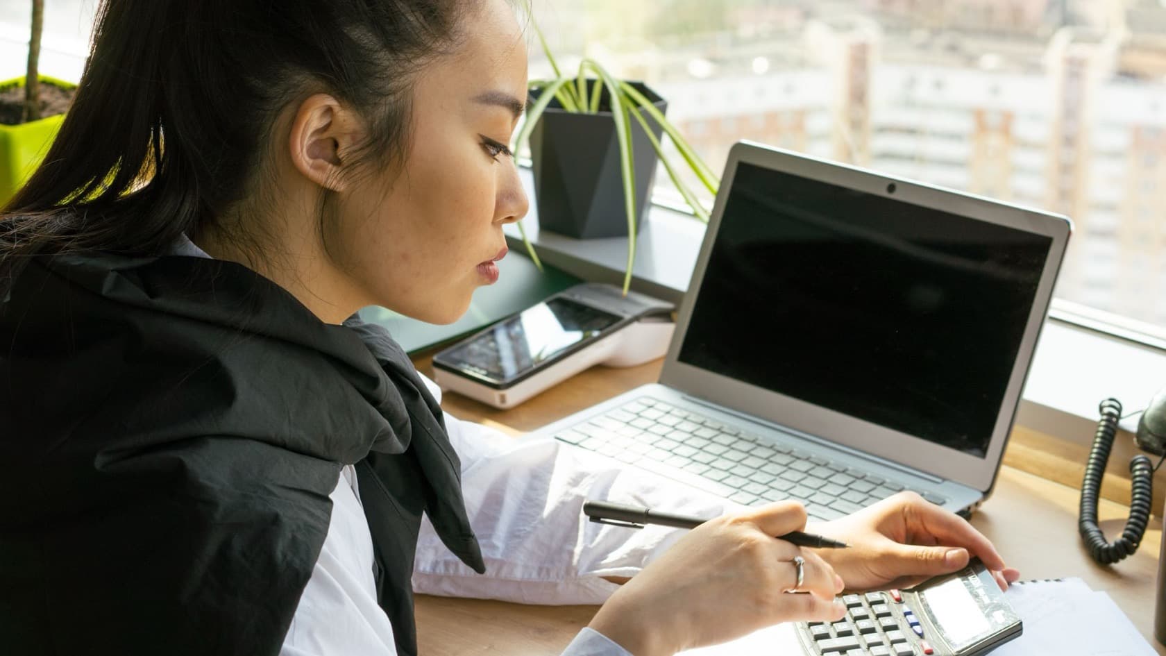 Office worker checking figures on a calculator beside an open laptop at a window desk