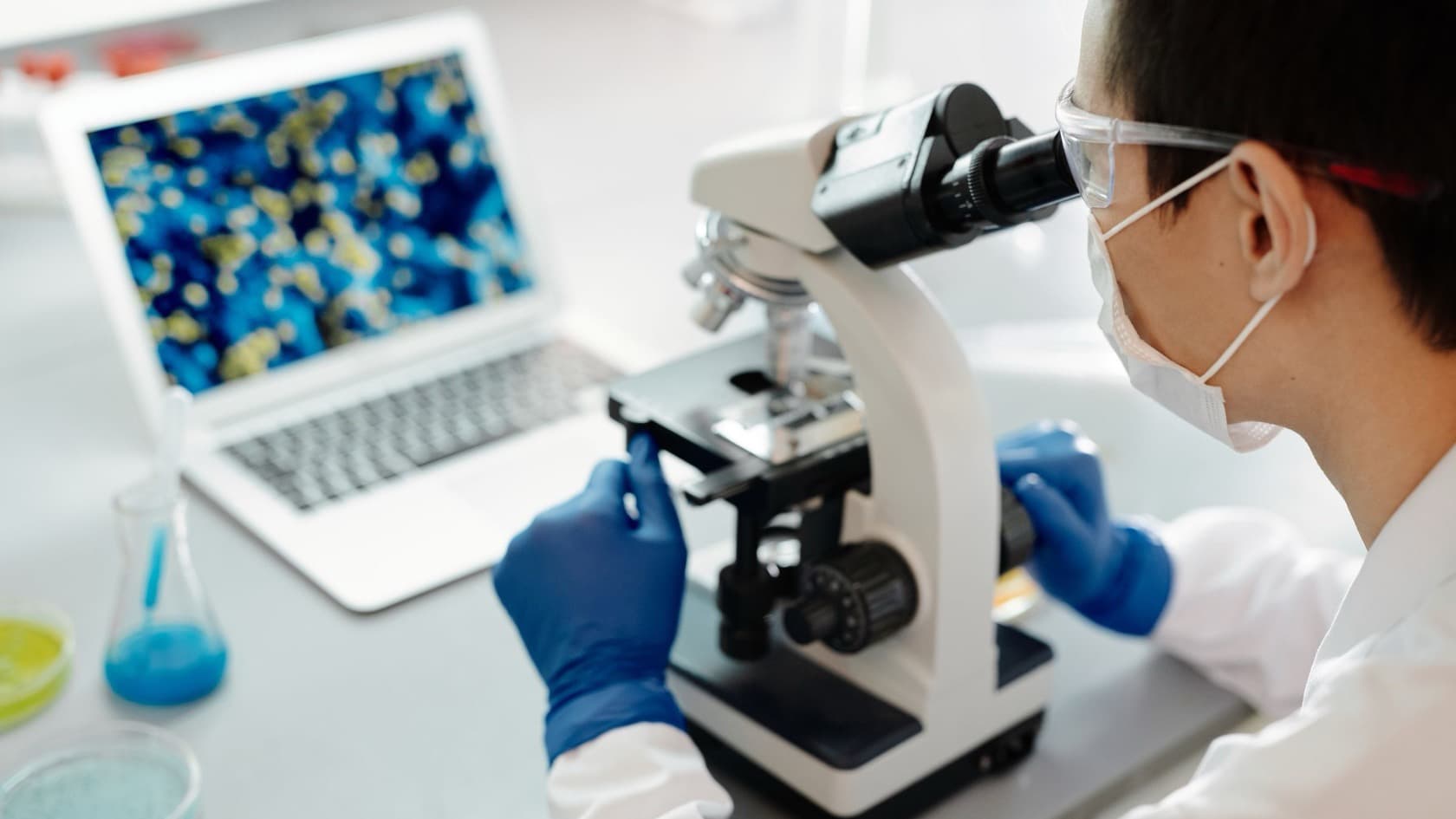 Scientist using a laboratory microscope beside a laptop showing a magnified sample