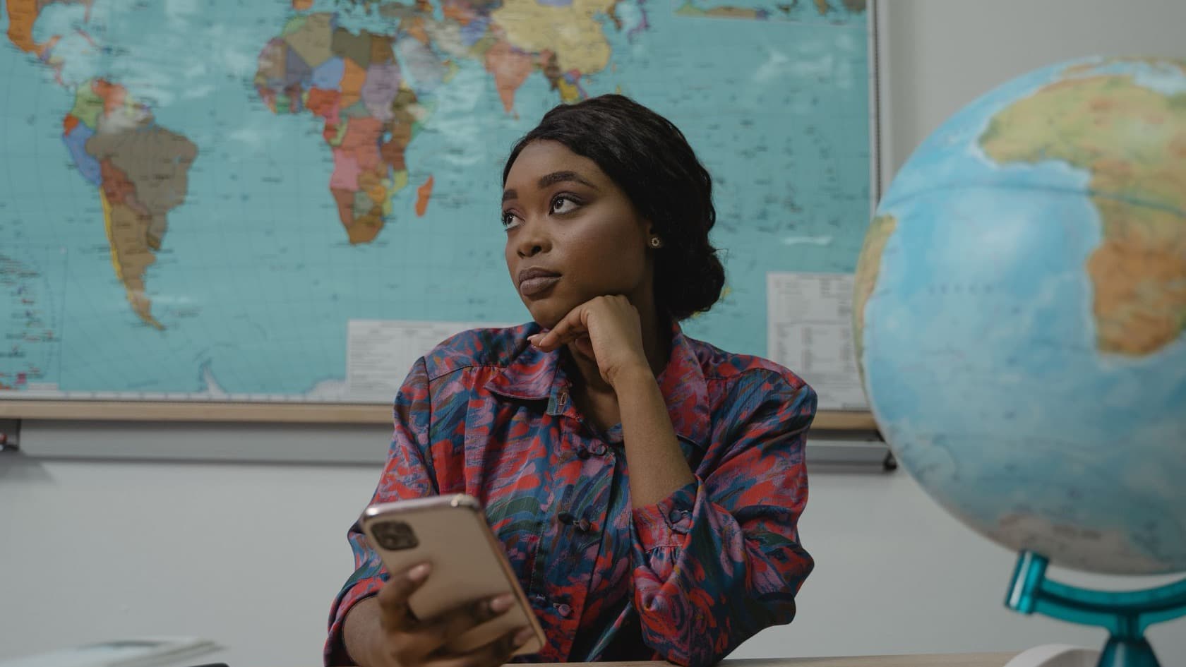 Teacher at her classroom desk holding a phone, world map and globe behind