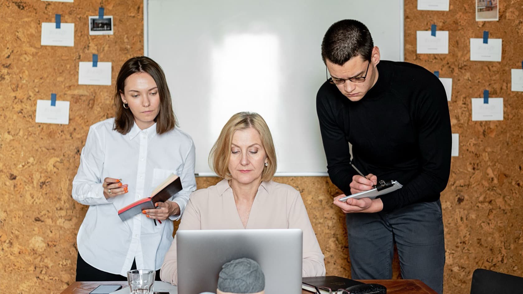 Three colleagues working through requests at a laptop, two taking notes, in front of a pinboard