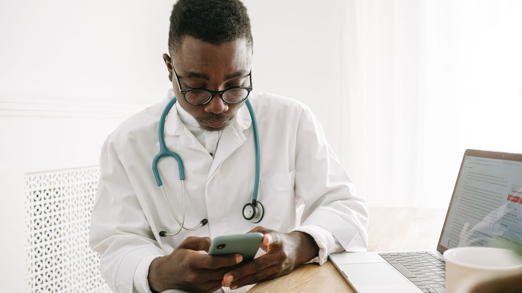 Doctor in a white coat using a phone at his desk beside an open laptop