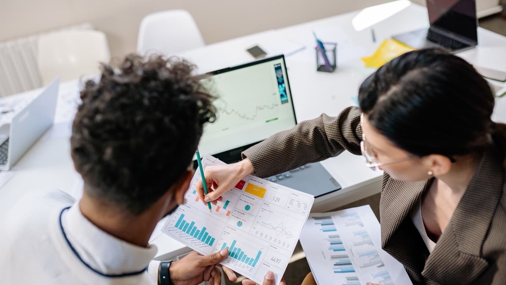 Two colleagues marking up printed charts beside a laptop dashboard