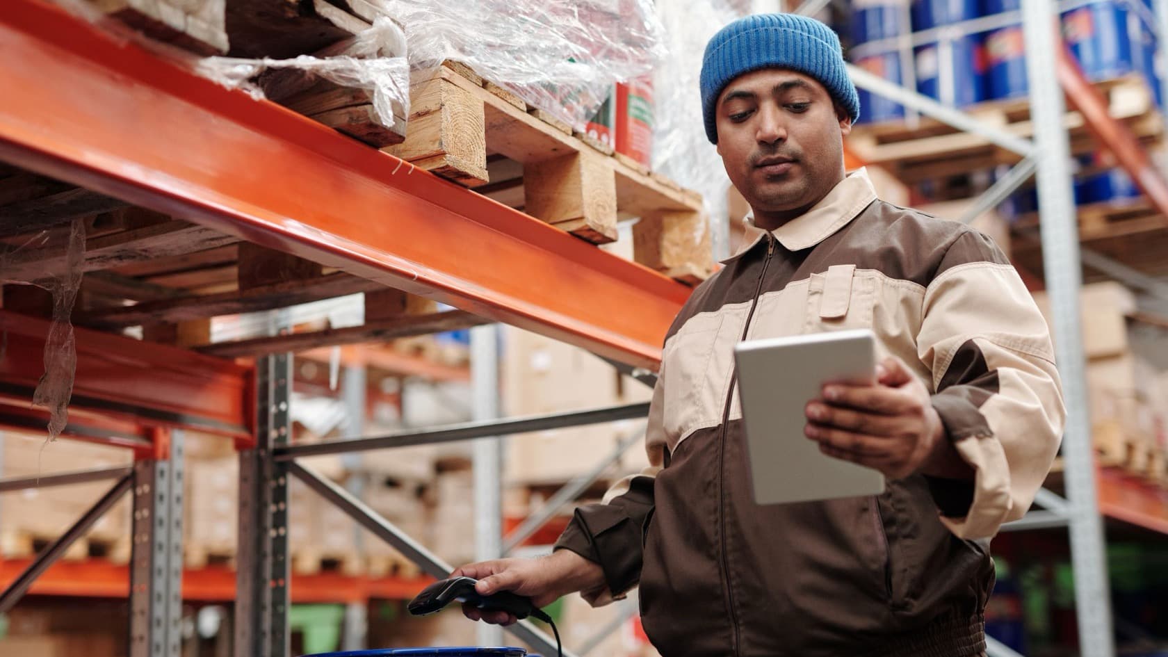 Warehouse worker checking stock on a tablet with a handheld scanner between red pallet racks