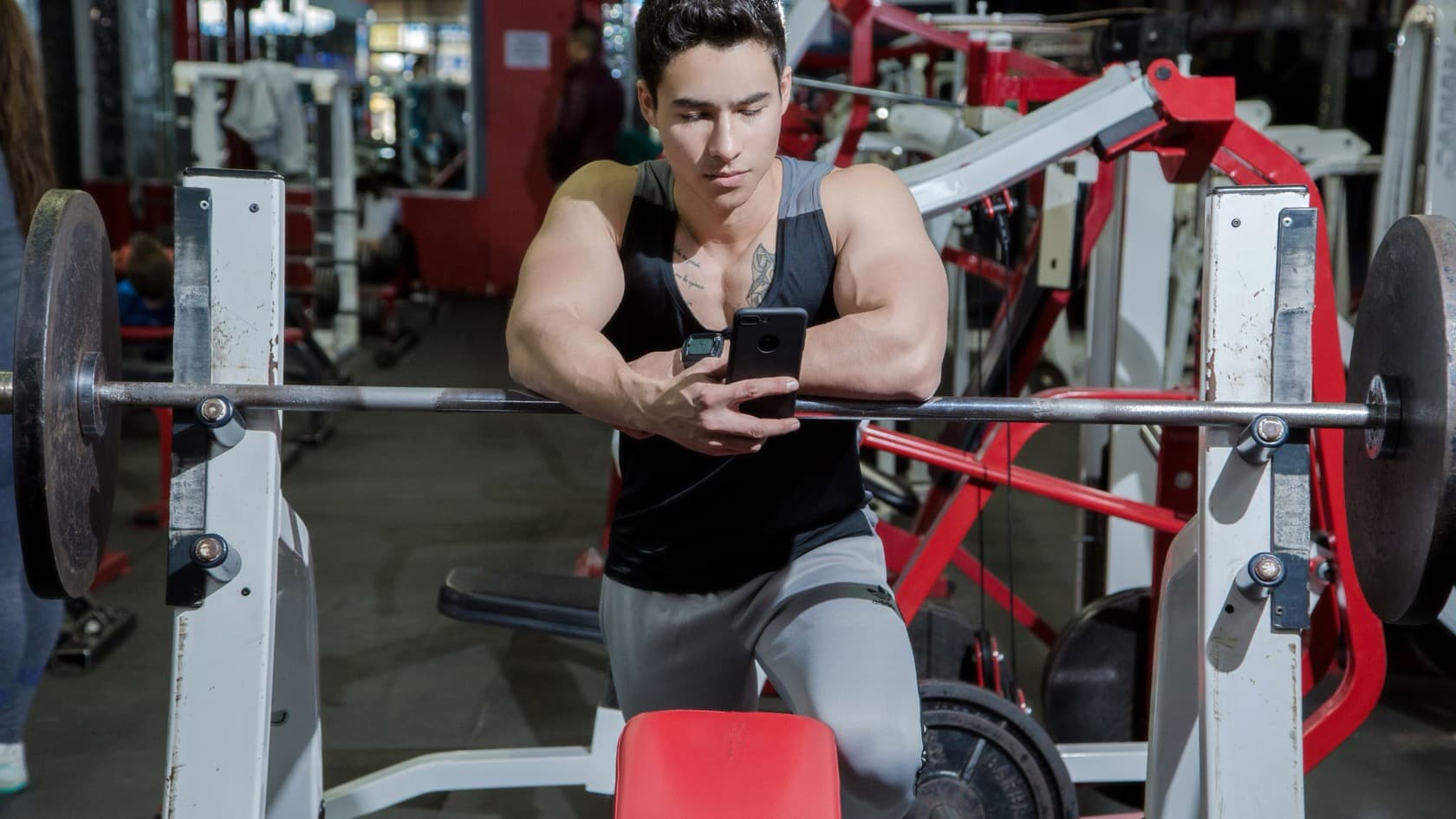 Gym member checking his phone while resting on a racked barbell, red machines behind