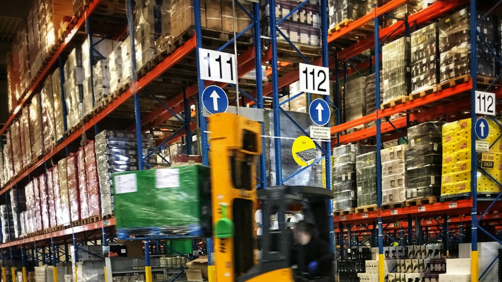 Forklift carrying a pallet between tall racks in a distribution warehouse